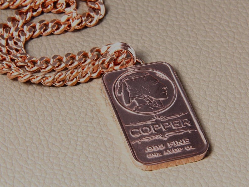 Puede incluir: Un collar de cadena de metal color cobre con un colgante rectangular en forma de lingote. El colgante muestra el perfil en relieve de una figura nativa americana, la palabra COPPER y el texto .999 FINE ONE AVDP OZ.