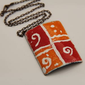 Puede incluir: Un collar con colgante rectangular que presenta una superficie de esmalte brillante y multicolor dividida en cuatro cuadrantes de rojo y naranja alternos. El diseño incluye patrones de espirales y puntos pintados a mano en blanco, sobre una cadena de metal oscuro.