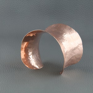 May include: A hammered copper cuff bracelet with a curved design. The bracelet is made from a single piece of copper that has been hammered to create a textured surface.