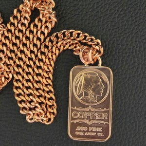 Puede incluir: Un collar de cadena de metal color cobre con un colgante rectangular. El colgante muestra el perfil en relieve de una figura nativa americana, la palabra COPPER y el texto .999 FINE ONE AVDP OZ. La cadena consta de eslabones entrelazados.