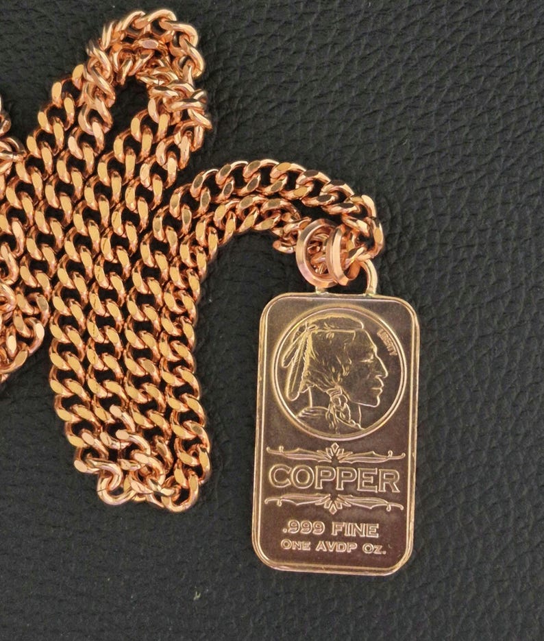Puede incluir: Un collar de cadena de metal color cobre con un colgante rectangular. El colgante muestra el perfil en relieve de una figura nativa americana, la palabra COPPER y el texto .999 FINE ONE AVDP OZ. La cadena consta de eslabones entrelazados.