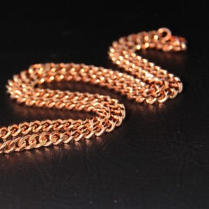 Puede incluir: Un collar de cadena de color cobre met&aacute;lico que presenta una serie de eslabones planos y ovalados entrelazados. La cadena est&aacute; dispuesta en una curva en forma de S, mostrando una superficie pulida y reflectante que capta la luz.