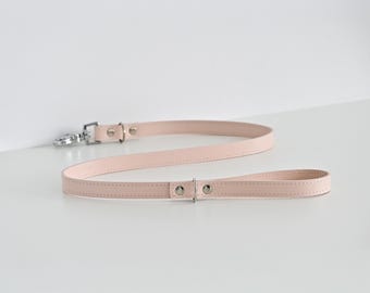 May include: A light pink leather dog leash with a matching handle. The leash has silver-colored metal hardware, including a clasp and rivets. The leash is approximately 48 inches long.