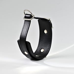 Pode incluir: Uma pulseira de couro preta com um anel e rebites de metal prateado. A pulseira é curva e tem uma borda costurada. O anel de metal é preso a um pedaço retangular de couro.