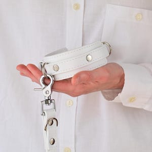 Könnte beinhalten: Ein weißes Lederhalsband- und Leinen-Set. Das Halsband ist gepolstert und mit silberfarbenen Metallbeschlägen befestigt. Die Leine hat einen passenden weißen Lederriemen und einen großen Karabinerhaken. Das Set wird in einer Hand vor einem weißen Hemd gehalten.