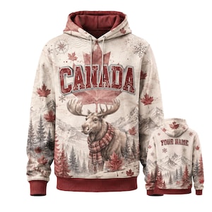 May include: A cream-colored hoodie featuring a large red maple leaf and the word CANADA in bold red letters. The design includes a moose wearing a plaid scarf, snowy mountain scenery, pine trees, and snowflakes. The back shows YOUR NAME text.