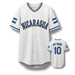 Puede incluir: Camiseta de béisbol blanca con "NICARAGUA" en letras azules en el pecho. La camiseta tiene detalles a rayas azules y blancas en las mangas y el cuello, y un parche de la bandera nicaragüense en las mangas. También se ve una camiseta más pequeña con "MY NAME" y el número "10".