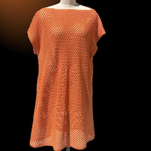 Może przedstawiać: Pomarańczowa sukienka zrobiona na szydełku, prezentowana na manekinie. Sukienka ma luźny, otwarty splot, dekolt w łódkę i krótkie rękawy. Sukienka sięga do kolan i ma lekko rozkloszowany dół. Tło jest czarne.