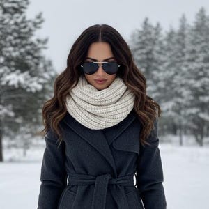 May include: A woman wearing a dark gray coat with a matching belt, a cream-colored knit scarf, and sunglasses. The background features a snowy landscape with evergreen trees, creating a winter scene.