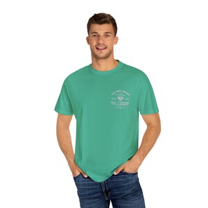May include: A teal green t-shirt with a small graphic on the upper left chest. The graphic includes text that reads "Cool Under Pressure" and "Tennis Club". The shirt is a solid color and has short sleeves.