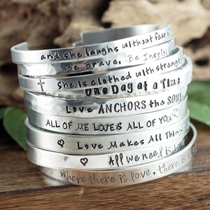 May include: A stack of silver bracelets with various inspirational messages engraved on them, including "and she laughs without fear", "Be Brave, Be Inspired", "She is clothed with strength", "One Day at a Time", "Love Anchors the Soul", "All of me loves all of you", "Love makes all things", "All we need is love", and "Where there is love, there is".