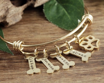 May include: Gold-colored bangle bracelet with four bone-shaped charms and a paw print charm. The bone charms are engraved with names: "Ozzee", "Connee", "Pu-kee", and "Bob-ee". The bracelet is displayed on a wooden surface.