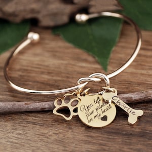 May include: Gold bangle bracelet with a knot design and charms. The charms include a paw print, a heart with the text "You left paw prints on my heart", and a bone with the name "Charlie" engraved on it.