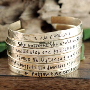 May include: A stack of gold-toned metal bracelets with various inspirational messages engraved on them, including "I am enough!" and "Follow your dreams".