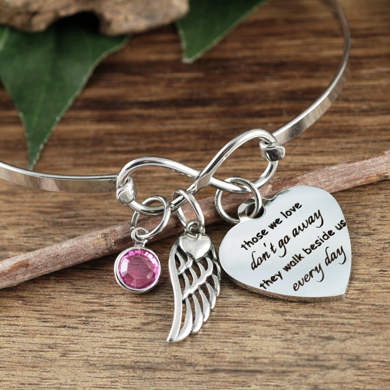 May include: Silver infinity bangle bracelet with a pink crystal charm, a silver heart charm, and a silver angel wing charm. The heart charm has the inscription "Those we love don't go away they walk beside us every day".