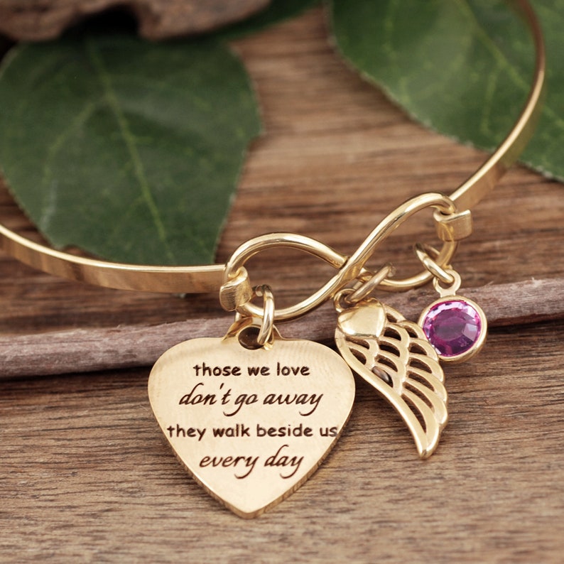 May include: Gold infinity bangle bracelet with a heart charm that reads "those we love don't go away they walk beside us every day" and a small angel wing charm with a pink crystal.