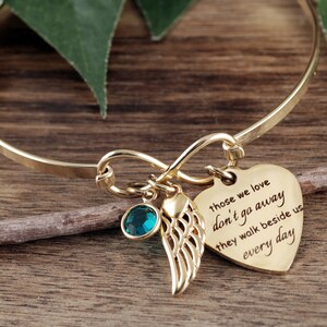 May include: Gold infinity bracelet with a heart charm that reads "those we love don't go away they walk beside us every day". The bracelet also features a small green crystal and a gold angel wing charm.