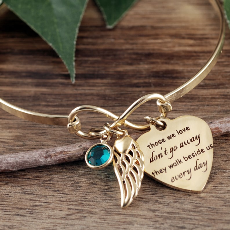 May include: Gold infinity bracelet with a heart charm that reads "those we love don't go away they walk beside us every day". The bracelet also features a small green crystal and a gold angel wing charm.