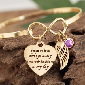 May include: Gold infinity bangle bracelet with a heart charm that reads "those we love don't go away they walk beside us every day" and a small angel wing charm with a purple gemstone.