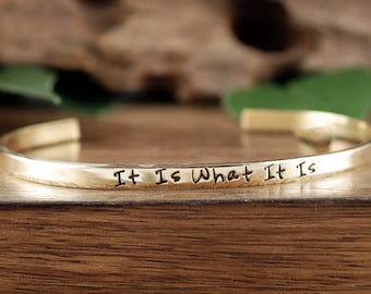 May include: A gold-toned bangle bracelet with the inscription "It Is What It Is" engraved on the front.