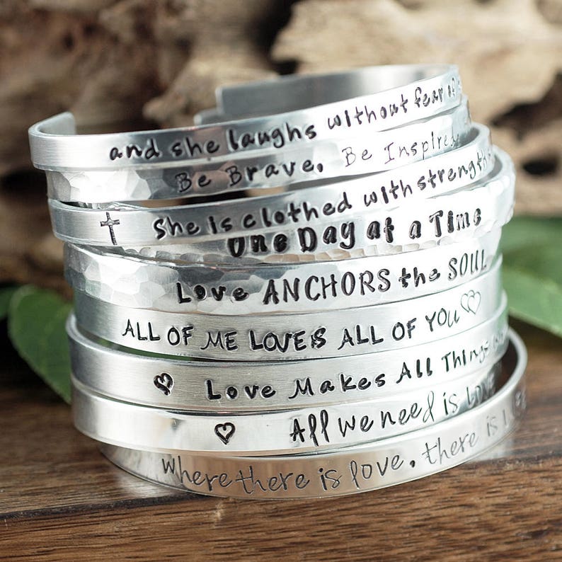May include: A stack of silver bracelets with various inspirational messages engraved on them, including "and she laughs without fear", "Be Brave, Be Inspired", "She is clothed with strength", "One Day at a Time", "Love Anchors the Soul", "All of me loves all of you", "Love makes all things", "All we need is love", and "Where there is love, there is".