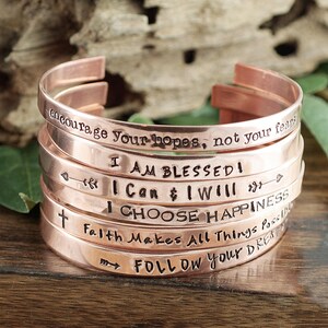 May include: A stack of six copper bracelets with inspirational messages engraved on them. The messages include "encourage your hopes, not your fears", "I am blessed", "I can & I will", "I choose happiness", "Faith makes all things possible", and "Follow your dreams".