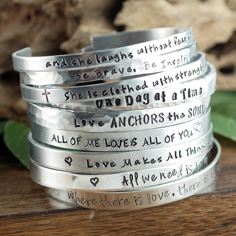 May include: A stack of silver bracelets with various inspirational quotes engraved on them. The quotes include "and she laughs without fear", "Be Brave", "She is clothed with strength", "One Day at a Time", "Love Anchors the Soul", "All of me loves all of you", "Love makes all things", "All we need is love", and "Where there is love, there is".