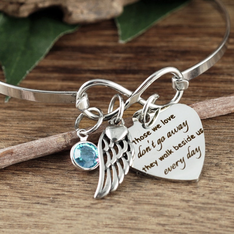 May include: Silver infinity bangle bracelet with a heart charm that reads "Those we love don't go away they walk beside us every day". The bracelet also features a small angel wing charm and a light blue crystal.