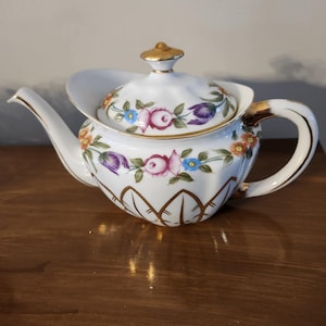 May include: White porcelain teapot with a floral design featuring pink, purple, and blue flowers. The teapot has a gold-colored handle, spout, and accents. The lid is topped with a gold finial. The teapot is sitting on a wooden surface.