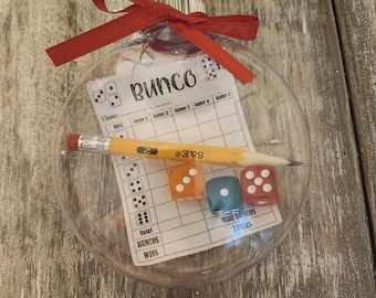May include: A clear spherical ornament containing a Bunco score sheet, a yellow pencil with an eraser, and three translucent dice in orange, blue, and red. The ornament is topped with a red ribbon bow tied around the silver metal cap at the top.