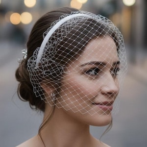 May include: A bride wearing a white birdcage veil with a delicate net design. The veil is attached to a white headband. The woman is wearing a strapless white dress. The background is a blurred street scene with soft lighting.