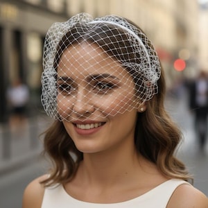 May include: A white birdcage veil attached to a thin headband. The veil features a classic diamond-shaped netting pattern that covers the upper portion of the face. The accessory is paired with a simple, sleeveless white dress with a round neckline.
