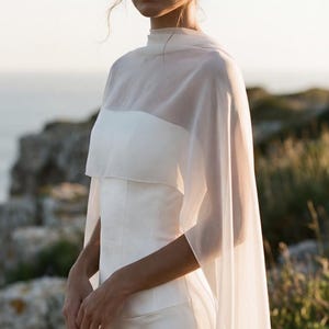 May include: A white wedding dress with a high neckline and a sheer cape. The dress has a fitted bodice and a flowing skirt. The cape drapes over the shoulders and arms. The background is a natural outdoor setting.