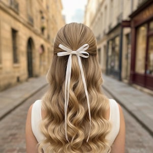 Könnte beinhalten: Blondes Haar, das mit einer weißen Schleife gebunden ist und in weichen Wellen fällt. Das Haar ist in einem Half-up-Half-down-Stil gestylt. Die Person trägt ein weißes Kleid. Der Hintergrund ist eine Kopfsteinpflasterstraße mit Gebäuden.