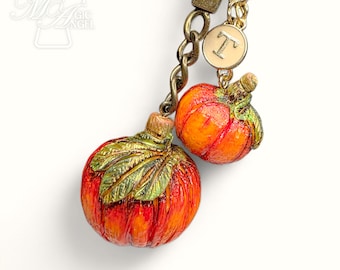May include: A fall-themed keychain featuring two miniature pumpkins. The pumpkins are painted in shades of orange and red with green leaves and stems. One pumpkin has a gold-colored initial charm. The keychain has a bronze-colored chain.