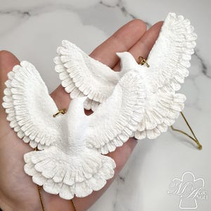 May include: Two white dove ornaments with outstretched wings. The doves have detailed feather textures and gold-colored hanging loops. The ornaments are likely for decoration or as a holiday accent.