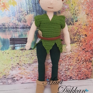May include: A handcrafted Peter Pan crochet doll, dressed in a green tunic, trousers, and hat with a red feather. The doll features brown hair and eyes, and tan boots. The backdrop depicts a park scene with a lake and autumn leaves.