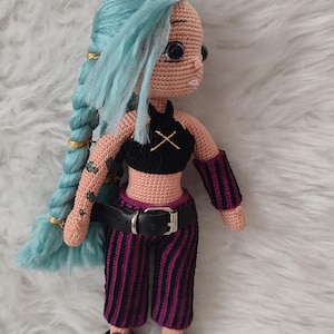 Puede incluir: Muñeca de ganchillo con cabello azul claro en una larga trenza, adornada con detalles dorados. La muñeca viste un top negro con diseño de X, cinturón negro y pantalones a rayas moradas y negras. Zapatos negros completan el atuendo.