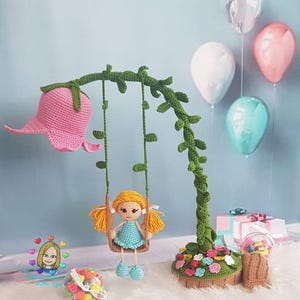Op de afbeelding: Een handgemaakte gehaakte pop op een schommel, met een roze bloem en groen wijnstokontwerp. De pop heeft geel haar en een blauwe jurk. De scène omvat kleurrijke ballonnen en een kleine geschenkdoos, alles op een zacht wit oppervlak.