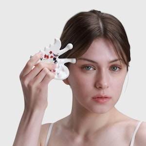 May include: A white ceramic facial massage tool with a butterfly-like shape. The tool features red and black floral accents. The person is holding the tool near their eye, suggesting its use for skincare or beauty treatments.