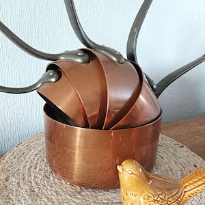 May include: A set of copper pots and pans with dark metal handles, nested together. A small, yellow ceramic bird figurine sits nearby on a woven mat. The copper cookware has a shiny, reflective surface.