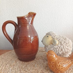 Puede incluir: Una jarra de cerámica marrón con asa, una figura decorativa de pájaro y una figura de oveja con cara color crema y lana esponjosa. Los objetos están dispuestos sobre una estera tejida.