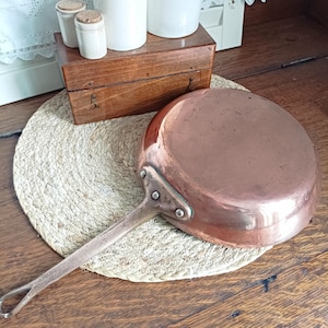 May include: A copper-colored frying pan with a long handle rests on a woven mat. A wooden box and several white ceramic jars with cork lids are in the background. The pan is round and has a riveted handle.
