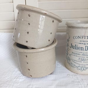 May include: Two stacked off-white ceramic cheese molds with small drainage holes along the sides, positioned next to a cylindrical off-white ceramic jar featuring black printed text that reads CONFIT, Julien D, MAISON PRINCIPALE, PARIS.