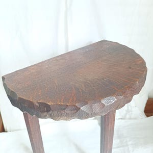 May include: A small, antique wooden table with a half-moon shaped top. The tabletop has a carved edge and is supported by three angled legs. The wood has a dark brown finish, showcasing the natural grain.