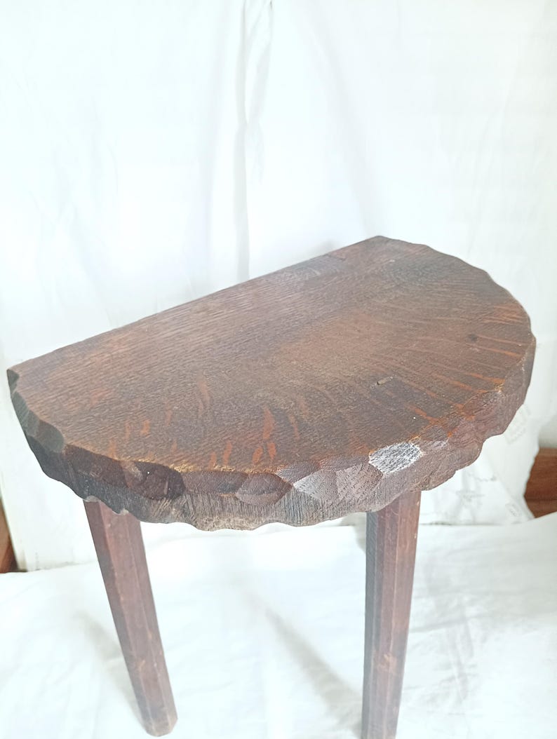 May include: A small, antique wooden table with a half-moon shaped top. The tabletop has a carved edge and is supported by three angled legs. The wood has a dark brown finish, showcasing the natural grain.