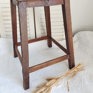 Peut inclure: Un tabouret en bois brun foncé avec une assise carrée dotée d'une petite découpe rectangulaire au centre. Le tabouret possède quatre pieds droits reliés par des barres de support horizontales sur les quatre côtés. Un bouquet de blé séché.
