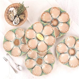 Peut inclure: Plusieurs assiettes à huîtres en forme de coquillage, chacune avec un puits central et des compartiments. Un petit panier d'huîtres et une branche d'herbes sont en arrière-plan. Une tranche de citron repose sur une assiette. Des couverts sont également présents.