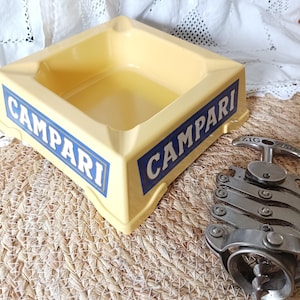 May include: A square, pale yellow ashtray featuring the blue and white text CAMPARI on two sides, accompanied by a metallic, accordion-style Zig-Zag corkscrew with a visible spiral worm and a hinged handle mechanism for opening wine bottles.