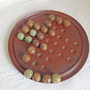 May include: A round wooden solitaire board game featuring a dark brown polished finish with numerous circular indentations. The board is populated with a collection of smooth, spherical marbles in various shades of brown, tan, and light green.
